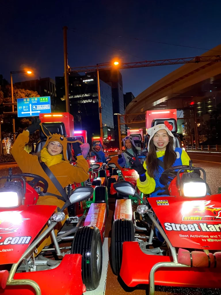 IMG_0022 - StreetKart Group of people in colorful costumes and animal hats posing on red go-karts at night on a city street with bright streetlights and buildings in the background, smiling at the camera.