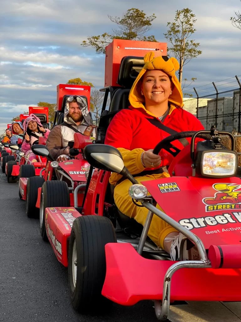 IMG_1123 - StreetKart Smiling woman in a yellow bear-hood onesie drives a red go‑kart at a costume parade, with costumed riders in line behind her.