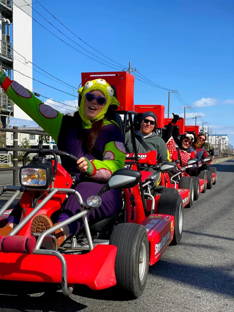 IMG_1486 - StreetKart Front rider in a bright green frog hat and purple costume waves from a red go-kart at a parade, others follow in line first-person view on a sunny street.