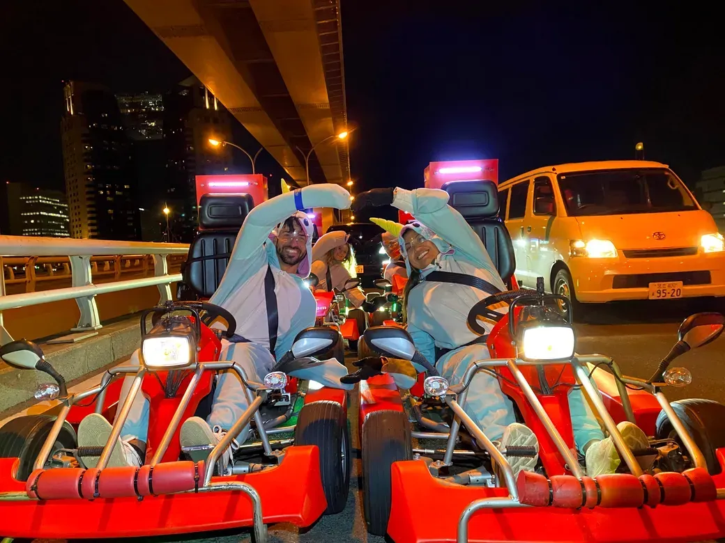Two people in white protective suits in red go-karts on a nighttime city road, forming a heart with their arms.